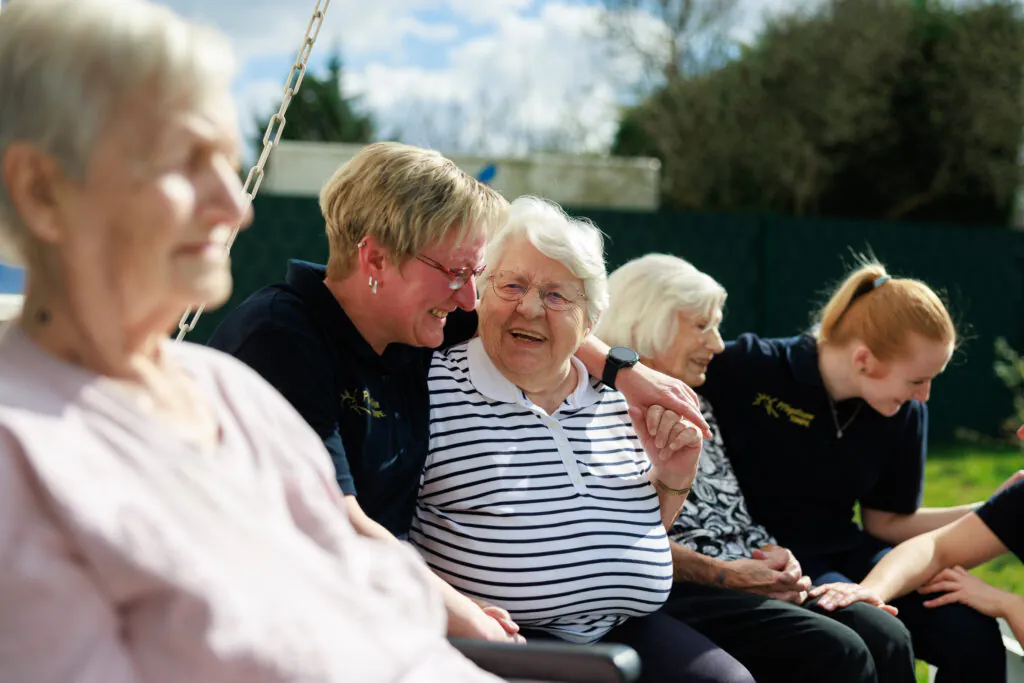 Seniorinnen und Pflegekräfte sitzen lachend zusammen auf einer Gartenschaukel