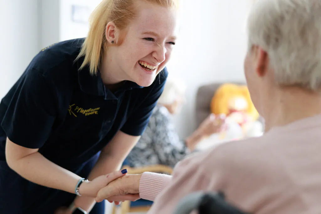Pflegekraft hält lachend die Hände einer Seniorin